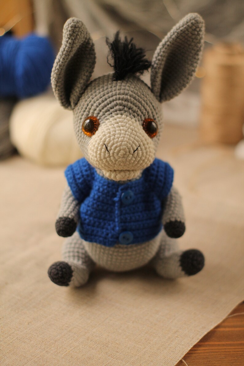 May include: A gray crocheted donkey wearing a blue crocheted vest with two buttons. The donkey has brown eyes and a black mane.