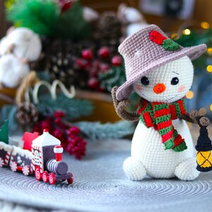 May include: A crocheted snowman wearing a brown hat with a red berry and green leaves, a green and red striped scarf, and holding a small black lantern. The snowman is sitting on a gray circular surface with a wooden toy train in the foreground.