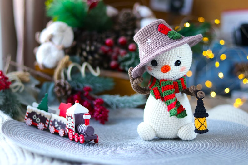 May include: A crocheted snowman wearing a brown hat with a red berry and green leaves, a green and red striped scarf, and holding a small black lantern. The snowman is sitting on a gray circular surface with a wooden toy train in the foreground.