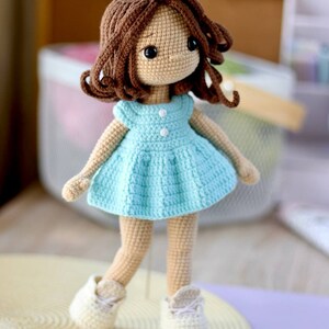 K&ouml;nnte beinhalten: Eine geh&auml;kelte Puppe mit braunen Haaren, einem hellblauen Kleid und wei&szlig;en Schuhen. Die Puppe steht auf einer hellbraunen Oberfl&auml;che.