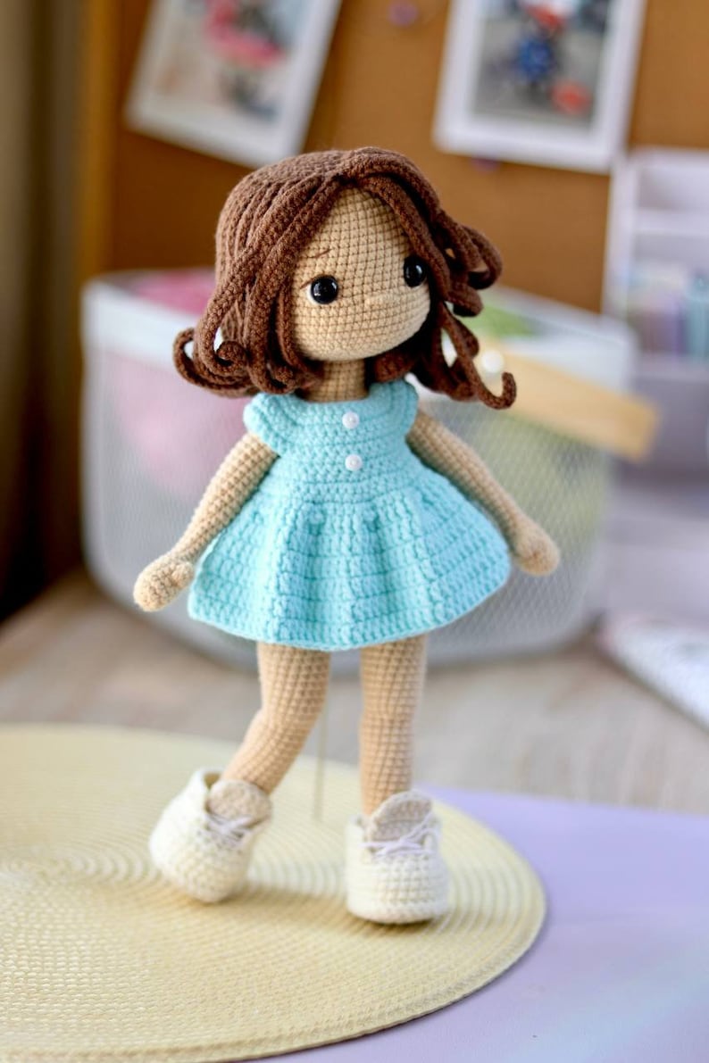 K&ouml;nnte beinhalten: Eine geh&auml;kelte Puppe mit braunen Haaren, einem hellblauen Kleid und wei&szlig;en Schuhen. Die Puppe steht auf einer hellbraunen Oberfl&auml;che.