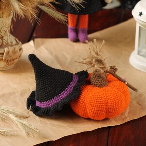 Peut inclure: Un chapeau de sorci&egrave;re en crochet avec une bordure violette et une citrouille orange en crochet. Le chapeau et la citrouille sont sur une surface en papier brun.