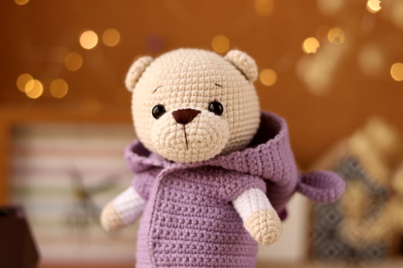 May include: A crocheted teddy bear wearing a purple hooded jacket. The bear has brown eyes and a brown nose.