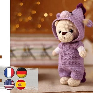 Op de afbeelding: Een gehaakte paarse teddybeer die een paarse outfit met capuchon draagt met een eenhoornhoorn. De beer heeft een wit gezicht en een bruine neus. De beer zit op een lichtbruin oppervlak.