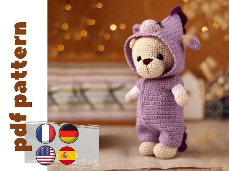May include: A crocheted purple teddy bear wearing a purple hooded outfit with a unicorn horn. The bear has a white face and brown nose. The bear is sitting on a light brown surface.