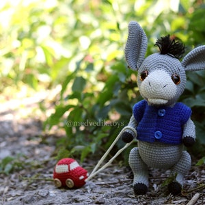 May include: A gray crocheted donkey toy wearing a blue crocheted vest with buttons, pulling a red toy car on a string. The donkey has brown eyes and a black mane. The toy is on a path with green foliage in the background.