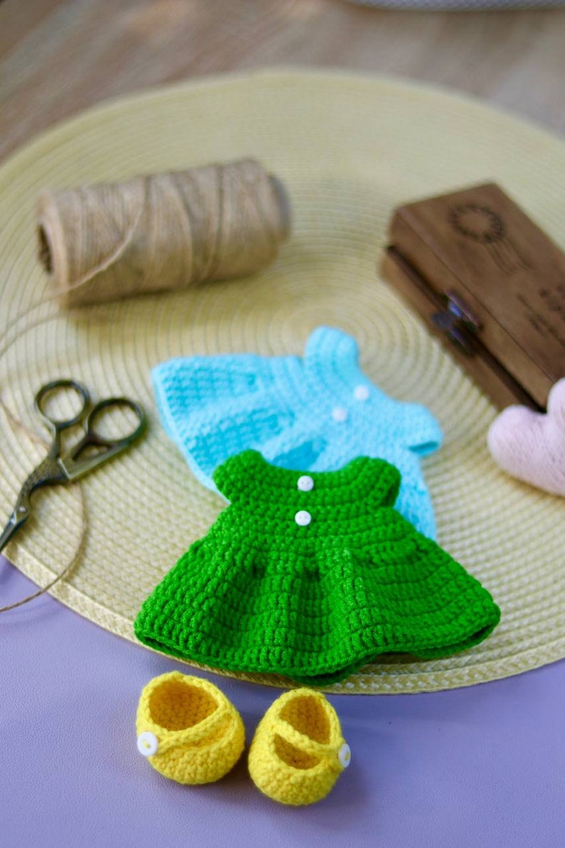 K&ouml;nnte beinhalten: Ein Set an geh&auml;kelten Puppenkleidern, bestehend aus einem gr&uuml;nen Kleid, einem hellblauen Kleid und einem Paar gelber Schuhe.