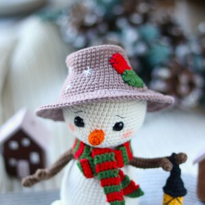 May include: A crocheted snowman wearing a brown hat with a red flower and green leaves, a red and green striped scarf, and holding a small lantern.