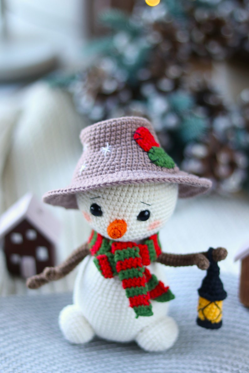 May include: A crocheted snowman wearing a brown hat with a red flower and green leaves, a red and green striped scarf, and holding a small lantern.