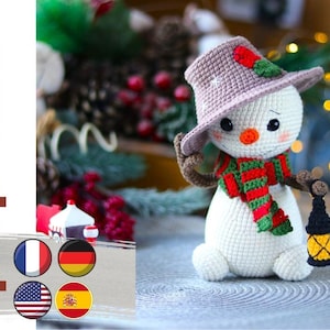 May include: Crocheted snowman amigurumi toy with a brown hat, red and green scarf, and a small lantern. The snowman is sitting on a gray surface with a blurry background of Christmas decorations.