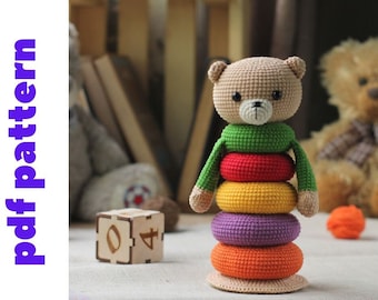 Könnte beinhalten: Ein gehäkeltes Teddybär-Spielzeug mit einem Stapel bunter Ringe. Der Bär ist braun mit einem grünen Shirt und die Ringe sind rot, gelb, orange, lila und orange.