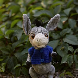 May include: A gray crocheted donkey wearing a blue crocheted vest with two blue buttons. The donkey has brown eyes and a black mane. The donkey is standing on a brown and gray surface with green foliage in the background. The text "@medvedik.toys" is visible in the lower right corner of the image.