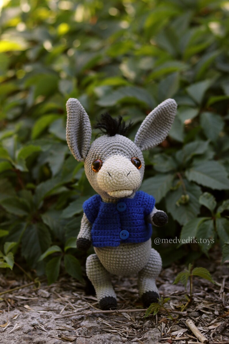 May include: A gray crocheted donkey wearing a blue crocheted vest with two blue buttons. The donkey has brown eyes and a black mane. The donkey is standing on a brown and gray surface with green foliage in the background. The text "@medvedik.toys" is visible in the lower right corner of the image.