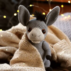 May include: A gray crocheted donkey toy with brown eyes and a black mane, wrapped in a cream-colored knitted blanket with gold glitter.