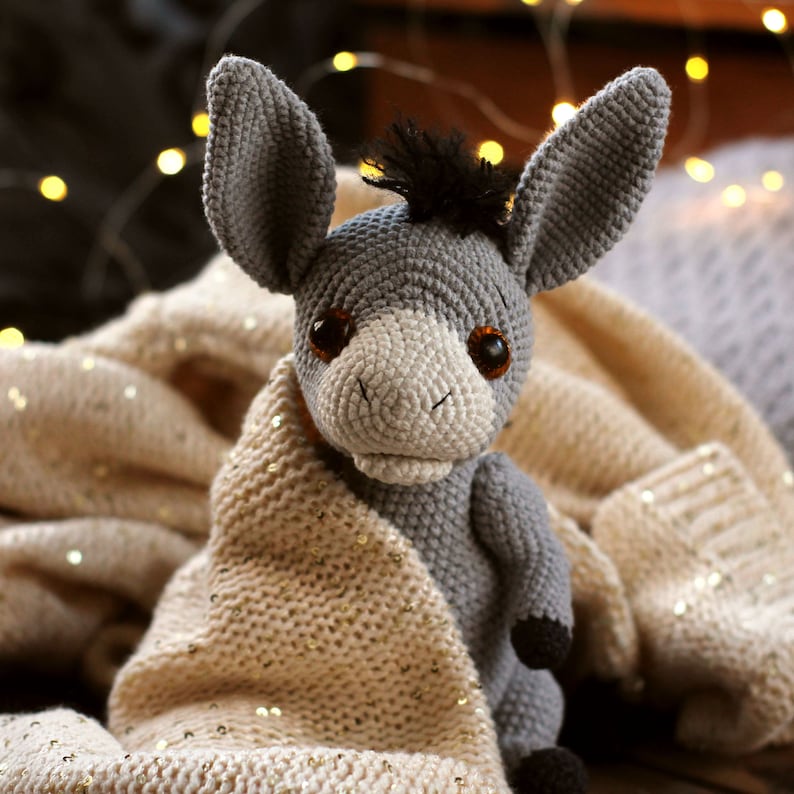 May include: A gray crocheted donkey toy with brown eyes and a black mane, wrapped in a cream-colored knitted blanket with gold glitter.
