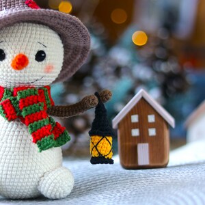 May include: A crocheted snowman wearing a brown hat and a red and green scarf holds a small yellow lantern. The snowman is standing on a white surface with a small wooden house in the background.