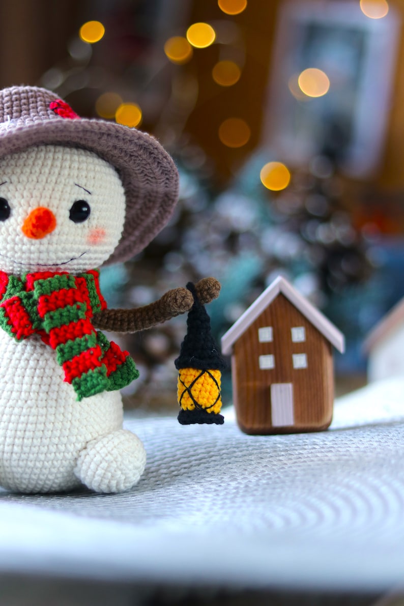 May include: A crocheted snowman wearing a brown hat and a red and green scarf holds a small yellow lantern. The snowman is standing on a white surface with a small wooden house in the background.