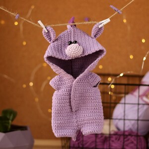 May include: A purple crocheted baby onesie with a hood shaped like a unicorn head. The onesie has a small, pink nose and black eyes.