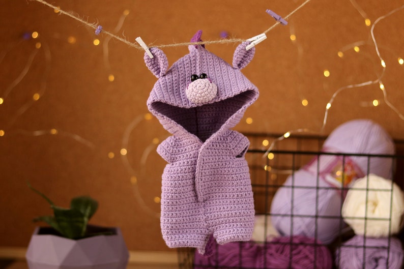 May include: A purple crocheted baby onesie with a hood shaped like a unicorn head. The onesie has a small, pink nose and black eyes.