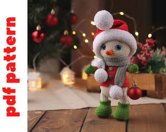 Könnte beinhalten: Ein gehäkeltes Schneemann-Spielzeug, das einen rot-weißen Weihnachtsmann-Hut, einen grauen Schal und grüne Stiefel trägt. Der Schneemann hält einen roten Weihnachtsbaumschmuck.