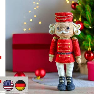 Könnte beinhalten: Gehäkelte Nussknacker-Puppe in roter Uniform mit goldenen Details, weißem Haar und weißem Schnurrbart. Die Puppe steht vor einem Weihnachtsbaum und einer roten Geschenkbox. Der Text "pdf pattern" ist auf der linken Seite zu sehen.