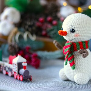 May include: A white crocheted snowman with a red and green striped scarf and a carrot nose. The snowman is sitting on a gray surface with a toy train in the foreground.