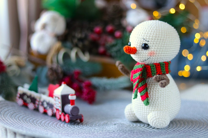 May include: A white crocheted snowman with a red and green striped scarf and a carrot nose. The snowman is sitting on a gray surface with a toy train in the foreground.