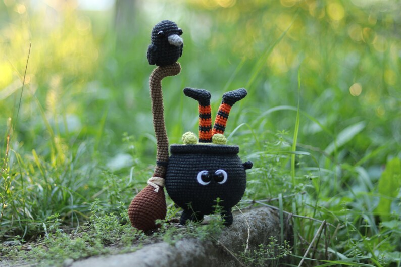 May include: A black crocheted cauldron with a witch's legs sticking out of the top. The cauldron has googly eyes and a brown crocheted broom.