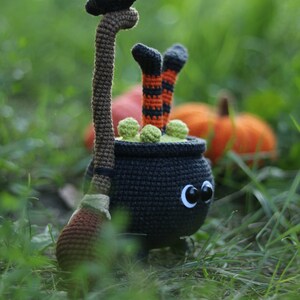 May include: A black crocheted cauldron with green goo and a black bird perched on a brown broomstick. The cauldron has large black eyes and is wearing orange and black striped stockings. The cauldron is sitting in a bed of green grass.
