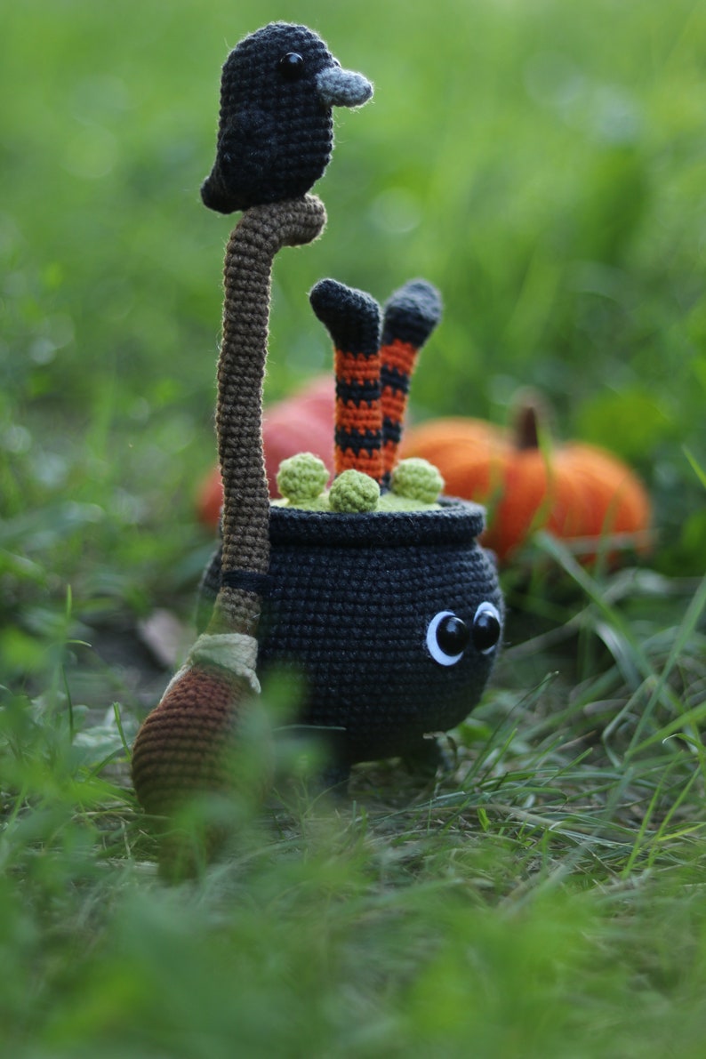 May include: A black crocheted cauldron with green goo and a black bird perched on a brown broomstick. The cauldron has large black eyes and is wearing orange and black striped stockings. The cauldron is sitting in a bed of green grass.