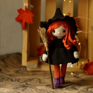 Peut inclure: Une poup&eacute;e de sorci&egrave;re en crochet portant un chapeau noir, des cheveux orange et une robe noire tenant un balai brun. La poup&eacute;e est debout sur une surface &agrave; motifs brun et blanc.