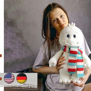 May include: A white crocheted plush toy with a red, white, and blue striped scarf. The toy has a smiling face with black eyes and a black mouth. The toy is being held by a person.