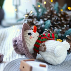 May include: A crocheted snowman toy with a brown hat, green and red striped scarf, and an orange nose. The snowman is lying on a white knitted blanket.