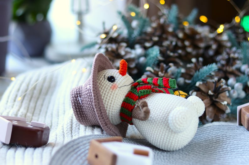 May include: A crocheted snowman toy with a brown hat, green and red striped scarf, and an orange nose. The snowman is lying on a white knitted blanket.