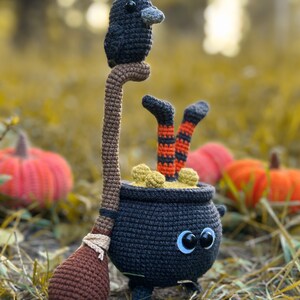 May include: A black crocheted cauldron with googly eyes and a brown crocheted broom. A black crocheted raven sits on top of the broom handle. The cauldron is filled with yellow crocheted ingredients. The cauldron is sitting on a bed of brown leaves.