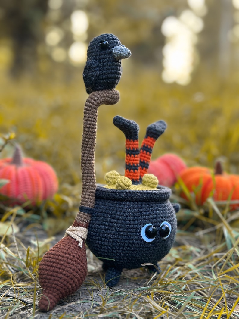 May include: A black crocheted cauldron with googly eyes and a brown crocheted broom. A black crocheted raven sits on top of the broom handle. The cauldron is filled with yellow crocheted ingredients. The cauldron is sitting on a bed of brown leaves.