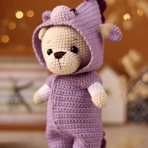 May include: A crocheted plush toy bear wearing a purple hooded onesie with a unicorn horn on the hood.