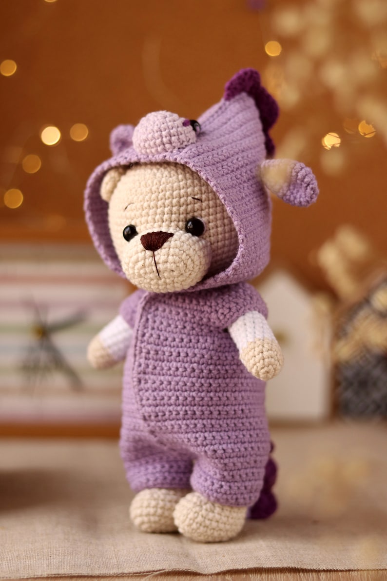 May include: A crocheted plush toy bear wearing a purple hooded onesie with a unicorn horn on the hood.