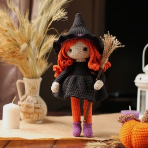Peut inclure: Une poup&eacute;e de sorci&egrave;re en crochet avec des cheveux orange, une robe noire et un chapeau violet tient un balai. La poup&eacute;e est debout sur une table en bois avec une bougie blanche, un vase avec des fleurs s&eacute;ch&eacute;es et une citrouille orange en crochet.
