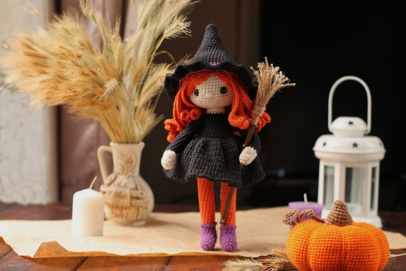 Peut inclure: Une poup&eacute;e de sorci&egrave;re en crochet avec des cheveux orange, une robe noire et un chapeau violet tient un balai. La poup&eacute;e est debout sur une table en bois avec une bougie blanche, un vase avec des fleurs s&eacute;ch&eacute;es et une citrouille orange en crochet.