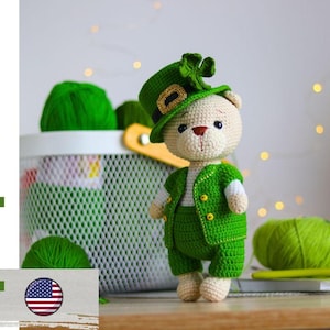 May include: Crocheted teddy bear wearing a green outfit and a leprechaun hat. The bear is standing on a wooden surface with a white wire basket and green yarn balls in the background. The text "pdf pattern" is on the left.