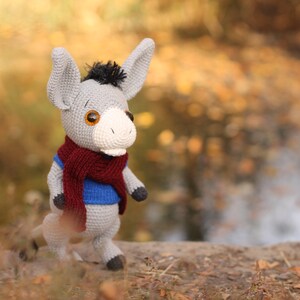 May include: A gray crocheted donkey wearing a blue sweater and a maroon scarf. The donkey is standing on a brown surface with a blurred background of fall foliage.
