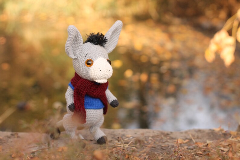 May include: A gray crocheted donkey wearing a blue sweater and a maroon scarf. The donkey is standing on a brown surface with a blurred background of fall foliage.