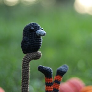 May include: A crocheted black bird with a grey beak perched on a brown stick. The bird is sitting on a green background with a blurry background.