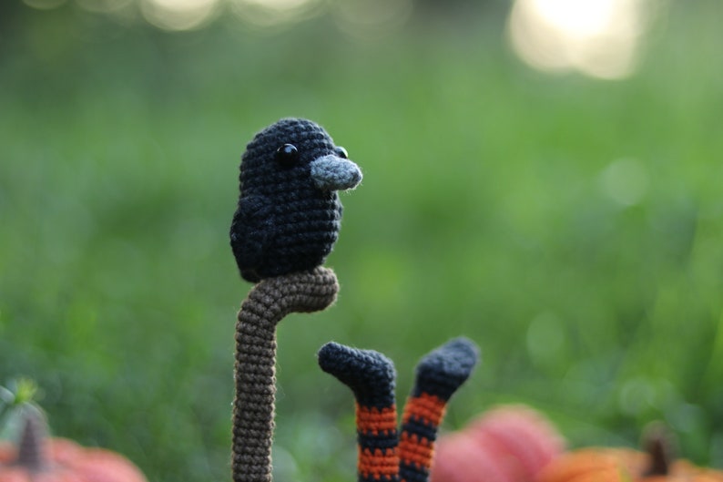 May include: A crocheted black bird with a grey beak perched on a brown stick. The bird is sitting on a green background with a blurry background.