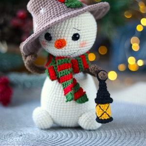 May include: A crocheted snowman wearing a brown hat with a red and green scarf and holding a small black lantern with a yellow window.