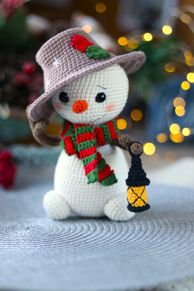 May include: A crocheted snowman wearing a brown hat with a red and green scarf and holding a small black lantern with a yellow window.