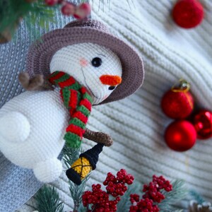 May include: A white crocheted snowman wearing a brown hat, red and green scarf, and holding a small lantern. The snowman is surrounded by red Christmas ornaments and a sprig of red berries.