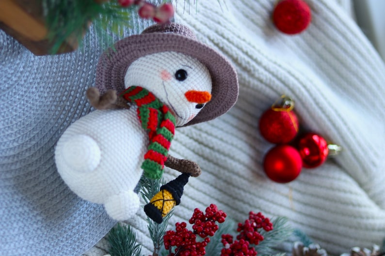 May include: A white crocheted snowman wearing a brown hat, red and green scarf, and holding a small lantern. The snowman is surrounded by red Christmas ornaments and a sprig of red berries.