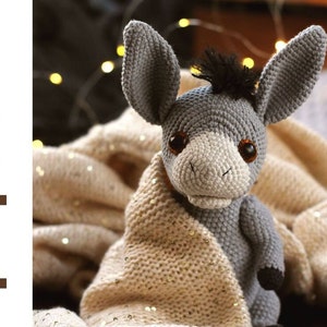 May include: A gray crocheted donkey toy with black yarn for the mane and tail. The donkey is sitting on a beige knitted blanket with gold glitter.